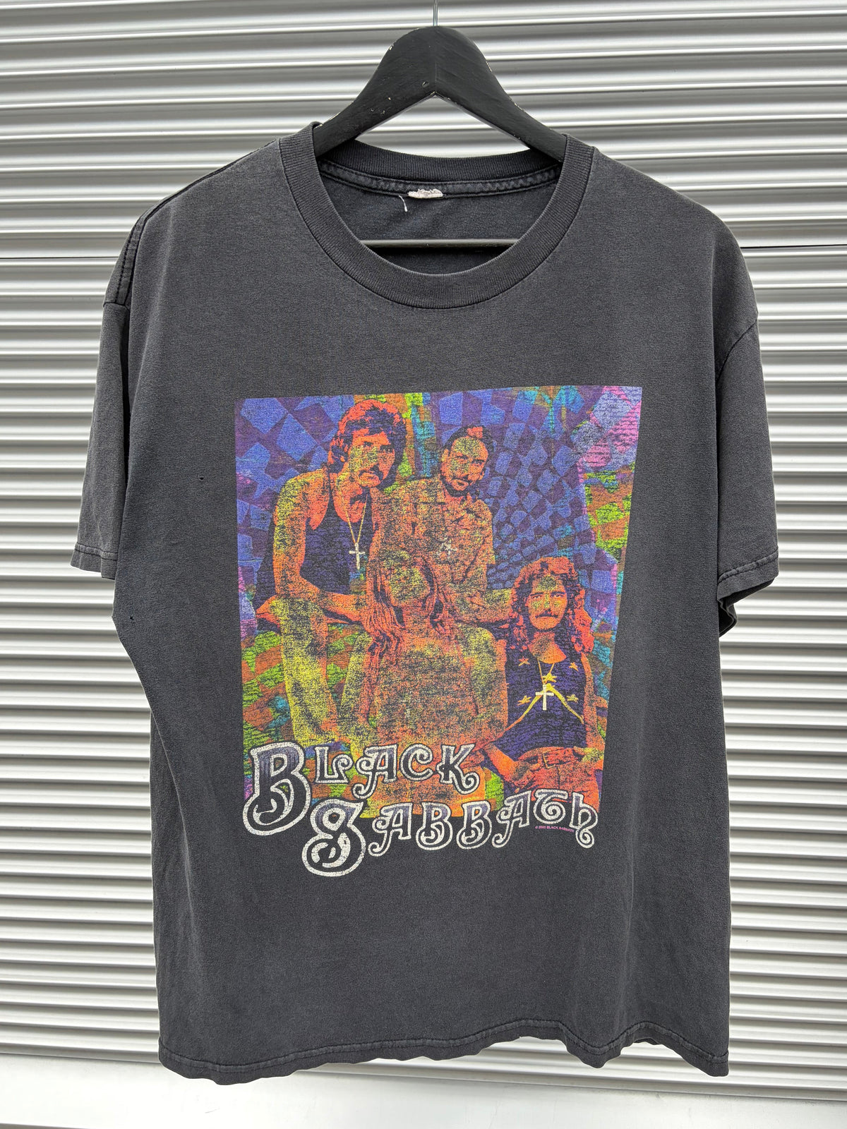 A faded black graphic t-shirt featuring the band Black Sabbath, laid flat on a gray corrugated metal background. The shirt has a large, colorful, psychedelic-style print of the band members on the front with the band's name in white gothic lettering.